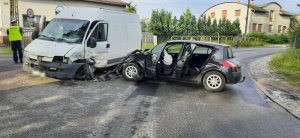 Na zdjęciu widać dwa rozbite samochody biorące udział w zdarzeniu. Auta zaparkowane na środku drogi. Z lewej strony zdjęcia widać policjanta.