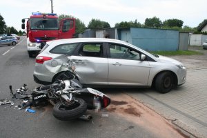 Na zdjęciu widać motocykl leżący na drodze i samochód osobowy, w który wjechał motocyklista. Za pojazdami widać wóz strażacki oraz policyjny radiowóz.
