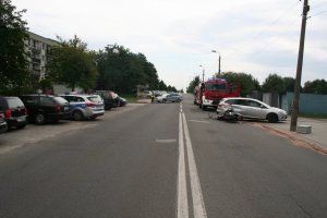 Z prawej strony zdjęcia widać motocykl leżący na drodze i samochód osobowy, w który wjechał motocyklista. Za pojazdami widać wóz strażacki oraz policyjny radiowóz, przy którym stoi policjantka. Z lewej strony zdjęcia widać zaparkowane przy drodze samochody.