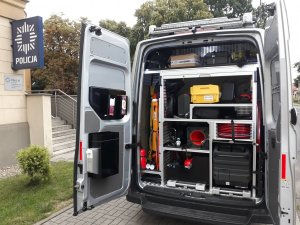 Zdjęcie kolorowe. Radiowóz oznakowany Volkswagen Crafter, który ma otwarte tylne drzwi i widoczne jest częściowo wyposażenie radiowozu. Auto zaparkowany jest przed tarnogórską komendą Policji.