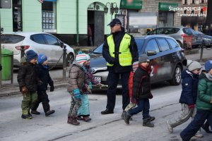 Na zdjęciu widać policjanta, który wstrzymał ruch samochodów, a przez przejście dla pieszych przechodzi grupa małych dzieci.
