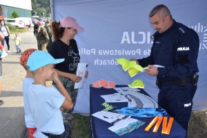 Na zdjęciu widać policjanta oraz matkę z dwójką synów. Policjant trzyma w ręku czapki z daszkiem promujące kampanię społeczną „Mamo! Tato! Tutaj jestem”.