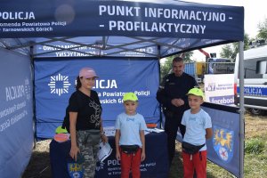 Na zdjęciu widać policjanta oraz matkę z dwójką synów. Dzieci mają na głowach czapki z daszkiem promujące kampanię społeczną „Mamo! Tato! Tutaj jestem”.