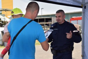 Na zdjęciu widać policjanta oraz mężczyznę z dzieckiem na ręku. Policjant rozmawia z rodzicem a dziecko ma na głowie  czapkę z daszkiem promującą kampanię społeczną „Mamo! Tato! Tutaj jestem”.
