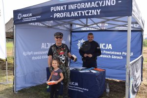 Na zdjęciu widać policjanta stojącego pod namiotem profilaktycznym. Przed nim znajduje się mężczyzna z dziewczynką, która otrzymała od policjanta gadżety.