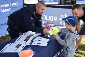 Na zdjęciu widać policjanta oraz ojca z synem. Policjant przekazał dziecku czapkę z daszkiem promującą kampanię społeczną „Mamo! Tato! Tutaj jestem”.