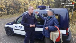 Na zdjęciu widać parking leśny, na którym przy radiowozie stoi policjant i rozmawia z grzybiarzem