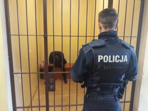Na zdjęciu widać policjanta, który obserwuje mężczyznę siedzącego za kratami-w policyjnej celi.