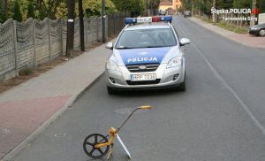 Na fotografii widać drogę, na środku której znajduje się policyjny radiowóz. Przed radiowozem, na jezdni widać przyrząd do pomiarów odległości.