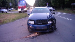 Na zdjęciu widać drogę w miejscu wypadku. Znajduje się na niej uszkodzony samochód skoda, w rowie samochód honda, a na poboczu widoczny jest wóz strażacki i policjanci wykonujący czynności. Na parkingu widać inne zaparkowane samochody.
