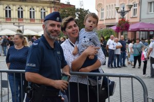 Na zdjęciu widać policjanta, obok którego stoi kobieta z dzieckiem na ręku. W tle zdjęcia widać innych uczestników imprezy.