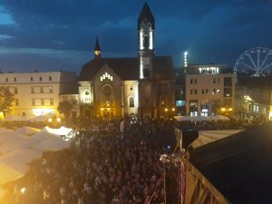 Zdjęcie przedstawia widok z góry na płytę rynku, na którym znajdują się uczestnicy imprezy, W tle zdjęcia widać oświetlone budynki i karuzele.