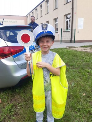 Na zdjęciu widać chłopca ubranego w kamizelkę odblaskową i policyjną czapkę. W ręku dziecko trzyma tarczę do zatrzymywania pojazdów. W tle zdjęcia widać radiowóz i policjanta.