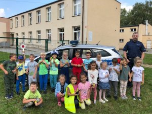 Na zdjęciu widać grupę dzieci stojących przy policyjnym radiowozie. W śród nich znajduje się policjant. Dzieci mają opaski odblaskowe, a jedno z nich, na głowie, policyjną czapkę.
