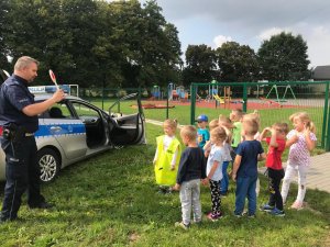 Na zdjęciu widać grupę dzieci stojących przed policjantem, który w dłoni trzyma tarczę do zatrzymywania pojazdów. Jedno z dzieci ma założoną odblaskową kamizelkę policyjną. W tle zdjęcia widać plac zabaw oraz policyjny radiowóz.