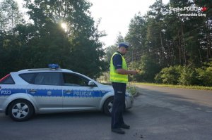 Na zdjęciu widać drogę biegnącą przez teren leśny. Przy drodze widać policjanta z Wydziału Ruchu Drogowego, który w dłoni trzyma urządzenie do badania stanu trzeźwości.