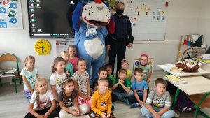 Zdjęcie przedstawia szkolną klasę, w której przy tablicy stoi dzielnicowy oraz maskotka Sznupek. przed policjantem, na podłodze siedzą uczniowie