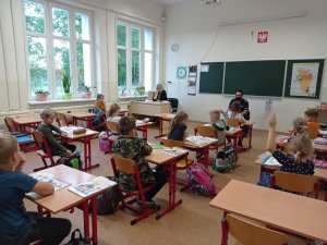 Zdjęcie przedstawia szkolną klasę. Przy tablicy siedzi na krześle policjant. Przed nim, w ławkach, siedzą uczniowie, a przy biurku nauczyciel.