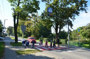 Na zdjęciu widać przedszkolaków przechodzących przez przejście dla pieszych. Przed przejściem stoi policjant, który zatrzymał samochód osobowy.