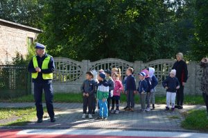 Policjant i nauczyciele wraz z grupą przedszkolaków stoją przed oznakowanym przejściem dla pieszych.