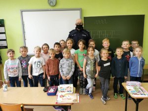Na zdjęciu widać szkolną klasę, w której pod tablicą, stoją uczniowie oraz dzielnicowy