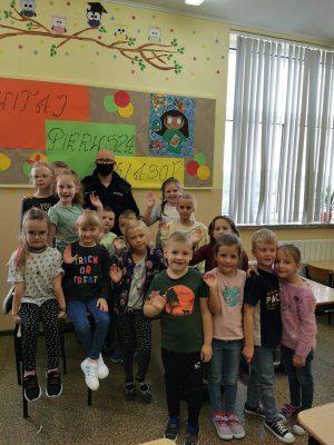 Na zdjęciu widać szkolną klasę, w której na ławce pod ścianą siedzą uczniowie oraz dzielnicowy. Na ścianie widać napis: Witaj pierwsza klaso!