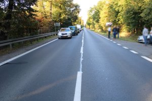 Zdjęcie przedstawia drogę. Z lewej strony widać trzy samochody biorące udział w zdarzeniu oraz zaparkowany za nimi policyjny radiowóz i policjanta kierującego ruchem. Z prawej strony widać policyjny radiowóz.