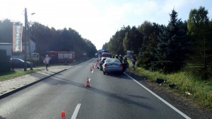 Na zdjęciu widać drogę prowadzącą przez teren leśny. Z prawej strony zdjęcia widać rozbite samochody biorące udział w zdarzeniu, policjantów i strażaków. Z lewej strony parking, na którym są zaparkowane samochody.