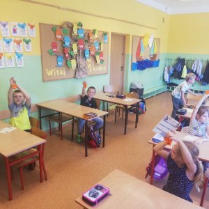 Szkolna klasa. W ławkach siedzą dzieci, które podnoszą ręce do góry, w których trzymają odblaski.