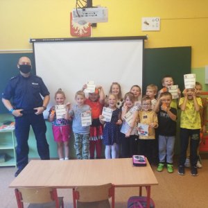 Szkolna klasa. Pod tablicą stoi dzielnicowy wraz z grupą dzieci. Dzieci trzymają w rękach kolorowanki i odblaski