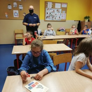 Szkolna klasa. W ławkach siedzą dzieci i oglądają odblaskowe gadżety. Pośród nich znajduje się dzielnicowy.