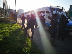 Grupa policjantów w hełmach stoi obok radiowozu. Policjanci obserwują stadion piłkarski