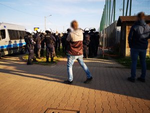 Grupa policjantów w hełmach stoi obok radiowozu. Policjanci obserwują stadion piłkarski