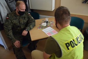 Przy stoliku siedzi policjant i żołnierz. Mundurowi wypełniają dokumenty