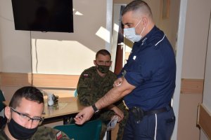 Policjant przykłada wacik do ręki w miejscu wkłucia igły. Przy stolikach siedzą żołnierze.