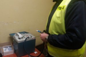 Policjant stoi obok urządzenia do badania stanu trzeźwości. W dłoni trzyma rurkę zakończoną ustnikiem.
