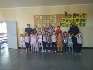 Na korytarzu szkoły, pod ścianą stoją policjanci i dzieci. Uczniowie trzymają w dłoniach odblaskowe elementy.