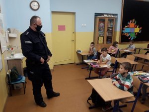 W szkolnej klasie policjant stoi obok tablicy i prowadzi pogadankę z uczniami. Dzieci siedzą w ławkach.