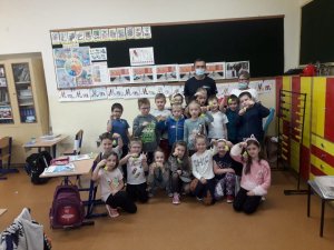 W szkolnej klasie, pod ścianą stoi policjant i dzieci. Uczniowie trzymają w dłoniach odblaskowe elementy.
