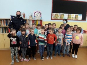 W szkolnej klasie, pod ścianą stoi policjant, nauczyciel i dzieci.