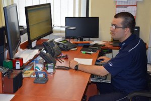 Na zdjęciu widać stanowisko pracy dyżurnego tarnogórskiej komendy. Policjant siedzi przy biurku i patrzy w monitor komputera. Na biurku widać telefony, radiostację oraz monitory komputerowe.
