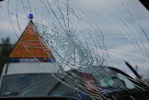 Rozbita szyba w samochodzie. W tle zdjęcia widać policyjny radiowóz, który na dachu ma trójkąt z napisem &quot;Wypadek&quot;