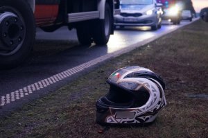 Na poboczu drogi, na trawie, leży kask motocyklisty. Na drodze widać policyjny radiowóz.