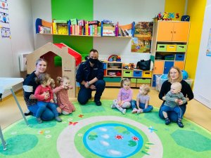 Sala szkolna. Na dywanie siedzą dzieci z opiekunami. Wśród nich znajduje się policjant