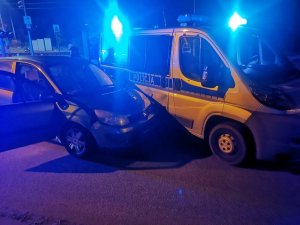 Na zdjęciu widać policyjny radiowóz z włączonymi sygnałami świetlnymi oraz samochód marki Renault Scenic, który wjechał w bok radiowozu.