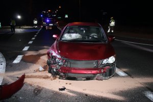 Na zdjęciu widać samochód, który ma uszkodzony przód. W tle zdjęcia widać strażaków oraz wóz strażacki