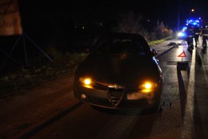 Na zdjęciu widać uszkodzony samochód osobowy w połowie zaparkowany na chodniku. Za pojazdem widać policyjny radiowóz i strażaków