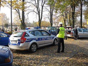Na zdjęciu widać policyjny radiowóz zaparkowany w rejonie cmentarza. Obok radiowozu stoi policjant obserwujący cmentarz