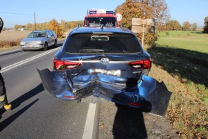 Na zdjęciu widać samochód osobowy, który ma zgnieciony tył. Przed samochodem znajduje się wóz strażacki, a obok drogą, jedzie inny samochód.