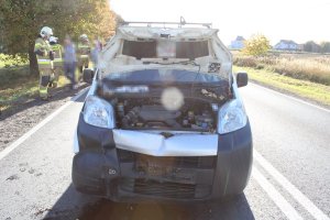 Na zdjęciu widać samochód osobowy, który ma uszkodzony przód. Auto ma podniesioną maskę - widać silnik. Za samochodem widać strażaków